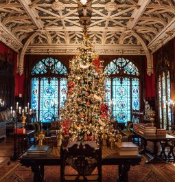 Beautifully decorated historic great room with large Christmas tree in center, dark brown wooden accents, detailed ceiling, and large stained-glass windows in background